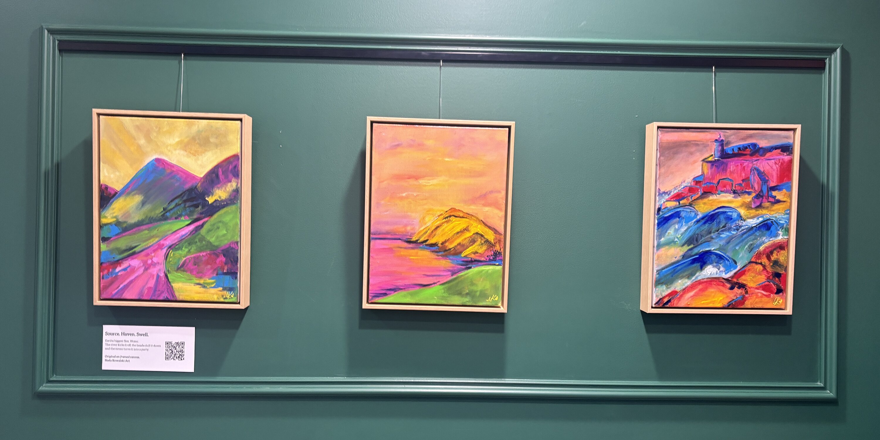 Three framed original paintings by Nada Kowalski hung side by side on a deep teal-green gallery wall, beneath a green picture-rail. From left to right: Source (abstract landscape in hot pink and blue), Haven (golden sunset mountain), and Swell (orange coastal scene).