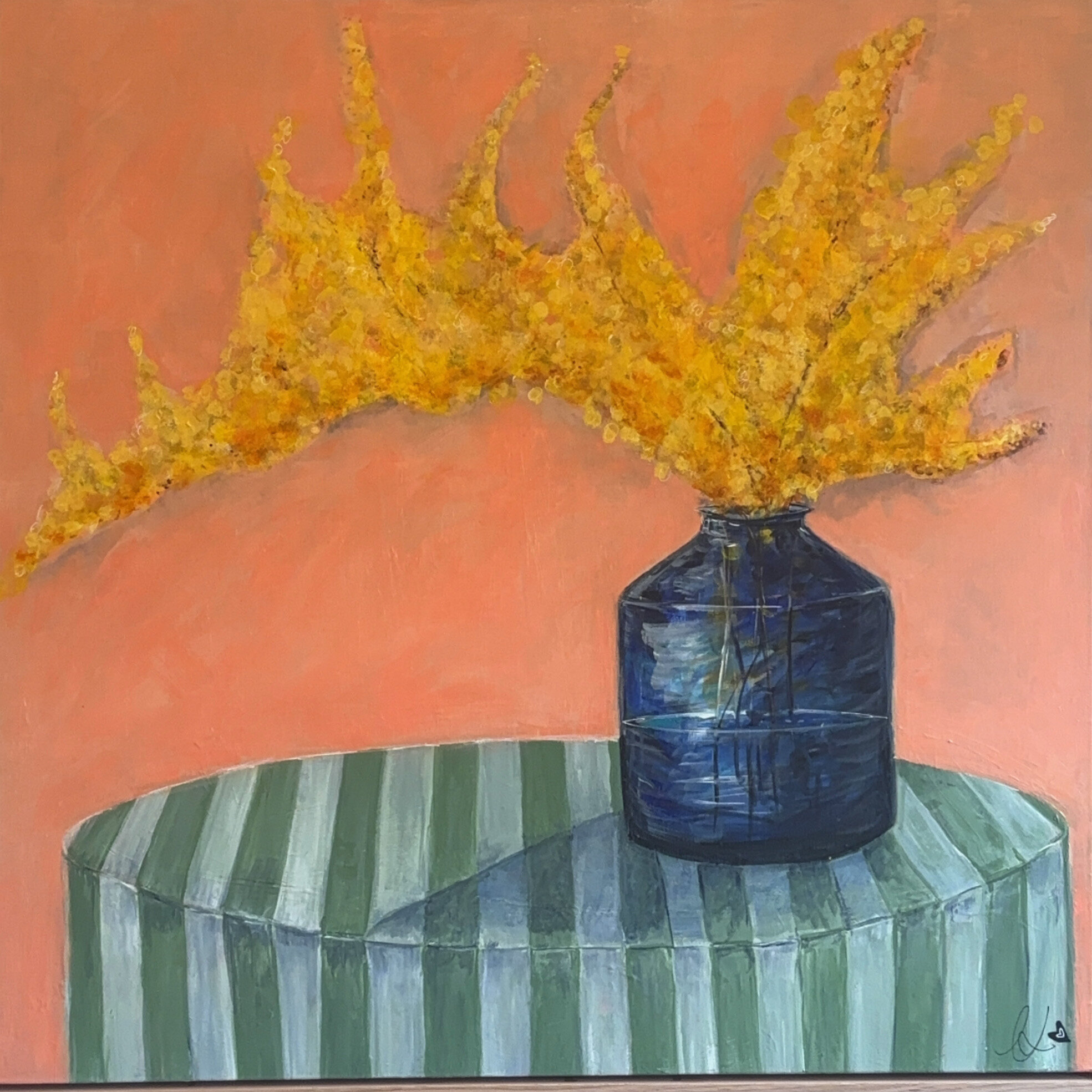Still life painting of a spray of bright yellow wattle blossoms in a cobalt blue glass bottle, sitting on a round green and white striped table against a coral peach background.